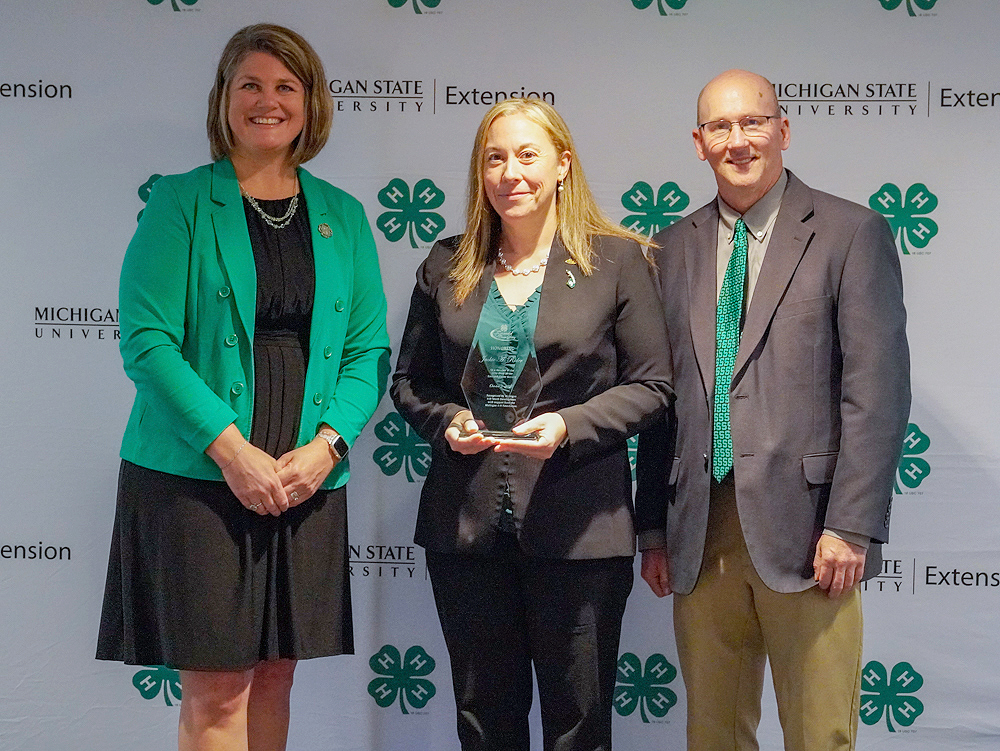 Three individuals at a backdrop. The center person holds an award.